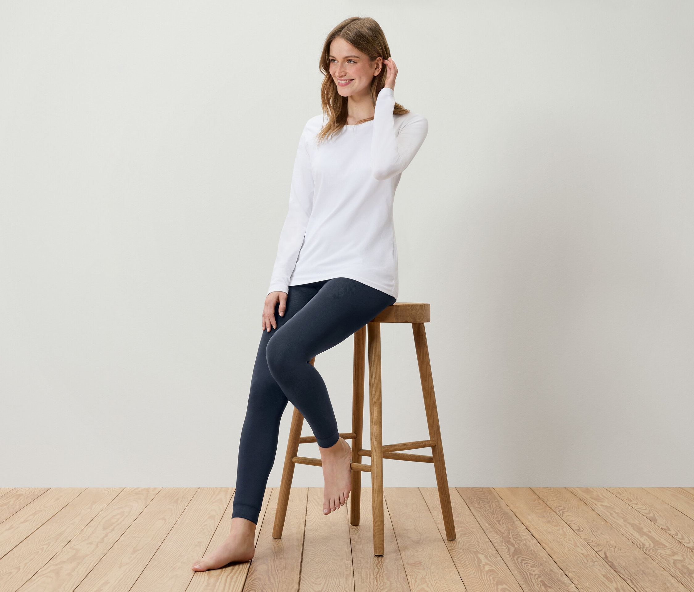 Mladá žena sedí na dřevěné stoličce, má na sobě bílé tričko s dlouhým rukávem a tmavě modré legíny.