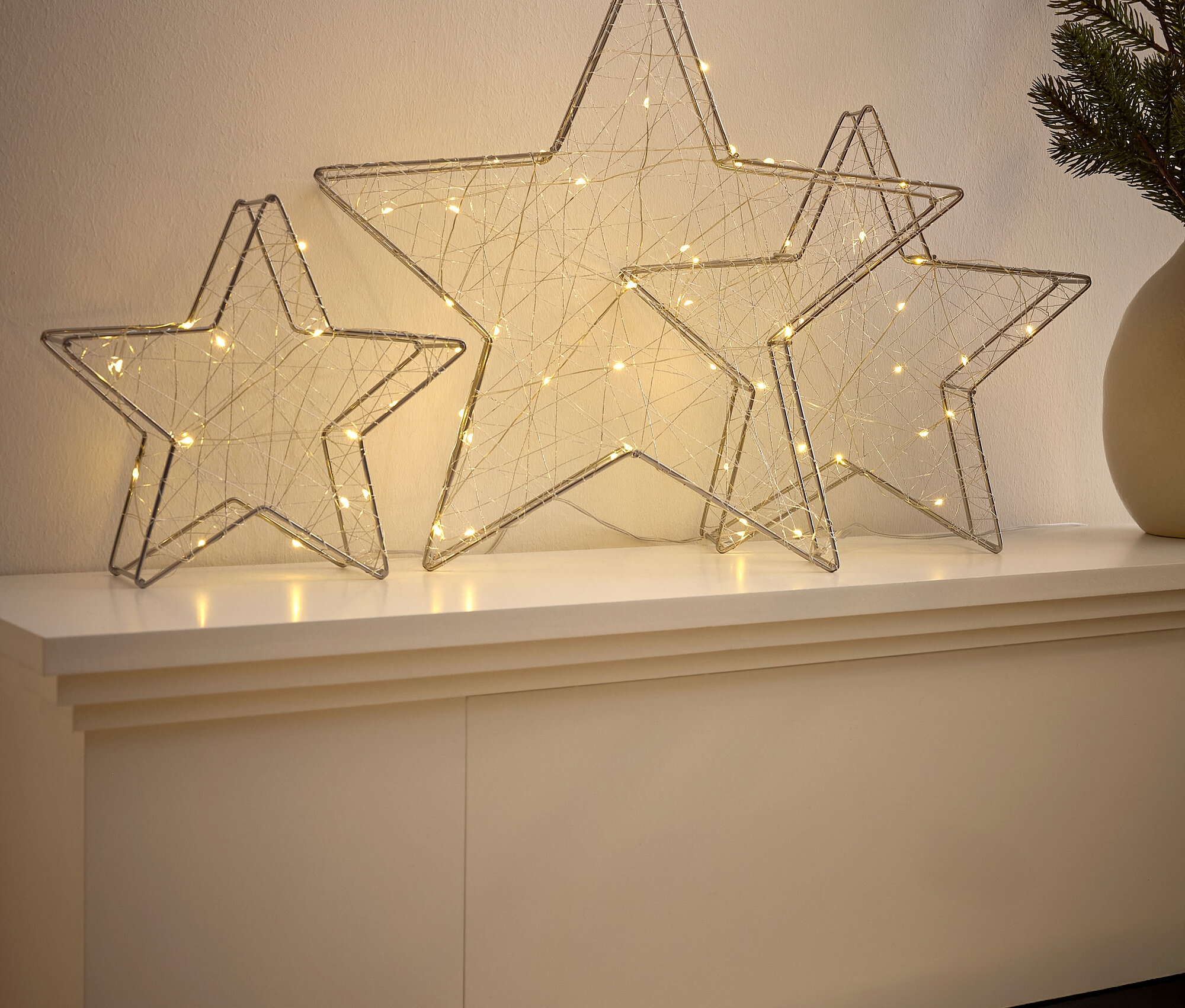 Tři drátěné hvězdy s bílými světýlky na krbu.