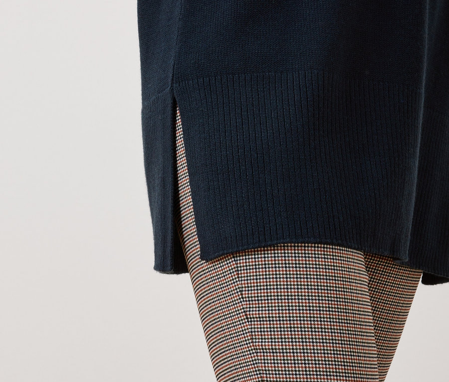 Detail dámského outfitu s tmavě modrým svetrem s rozparkem a kostkovanými kalhotami.