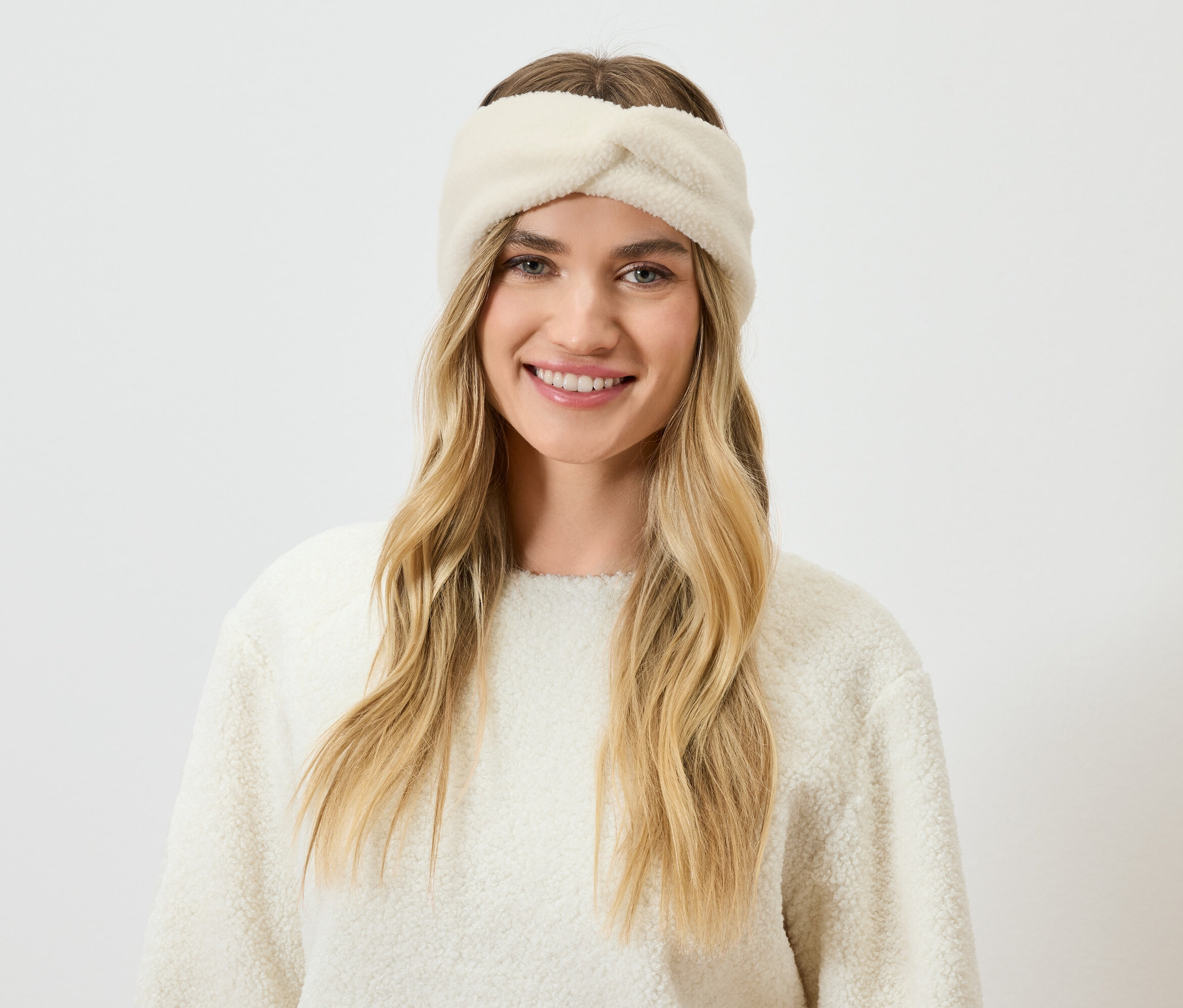 Portrét usměvavé ženy s blond vlasy a krémovou čelenkou a svetrem.