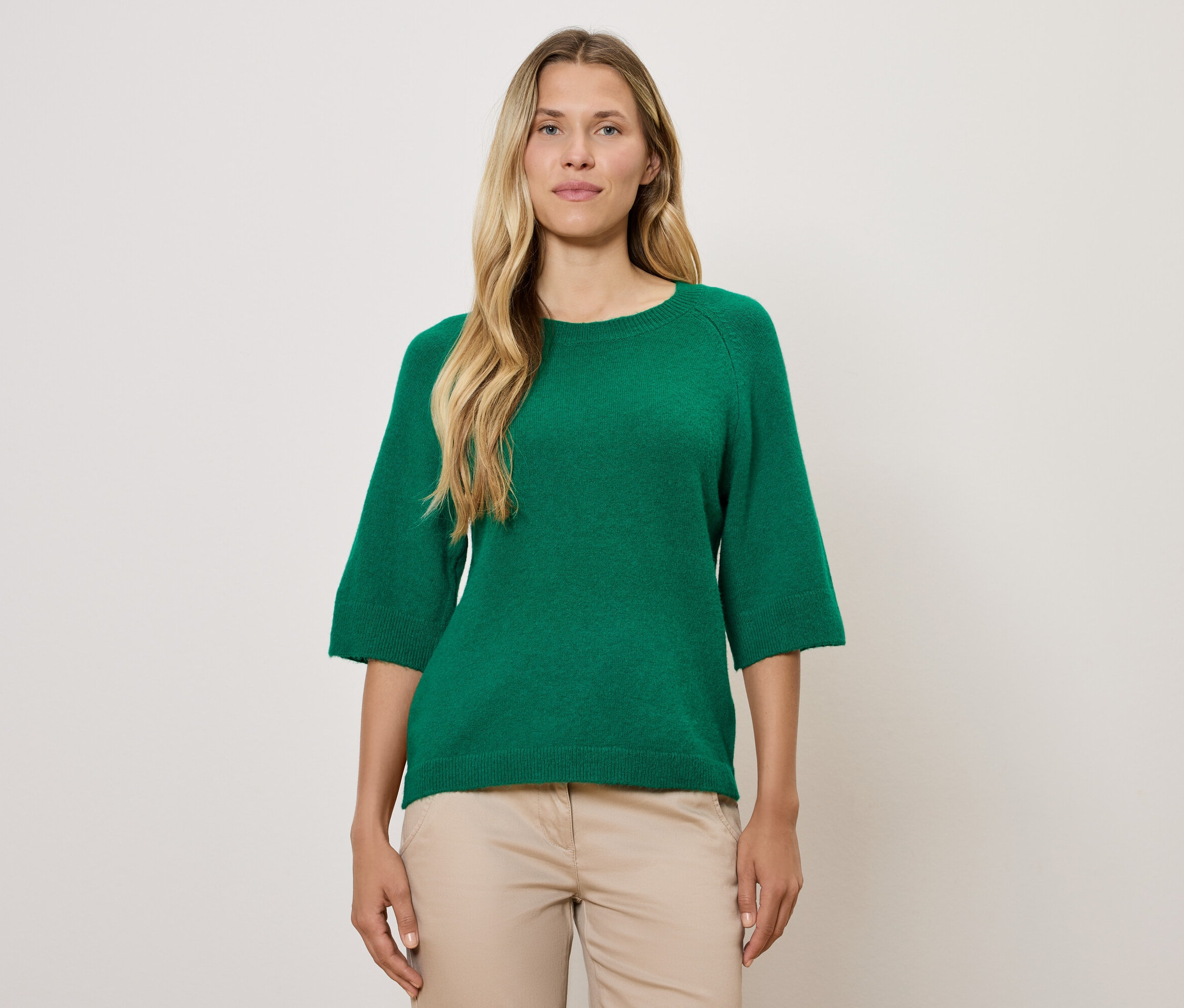 Blondýna má na sobě zelený svetr s tříčtvrtečními rukávy a béžové kalhoty.