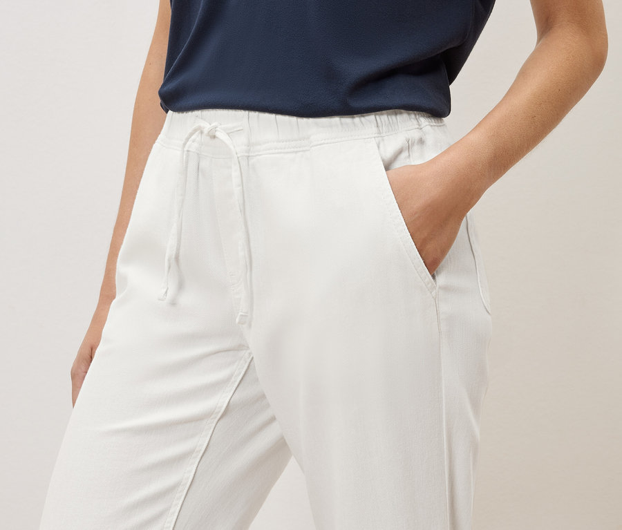 Žena má ruku v kapse bílých kalhot s elastickým pasem a šňůrkou.