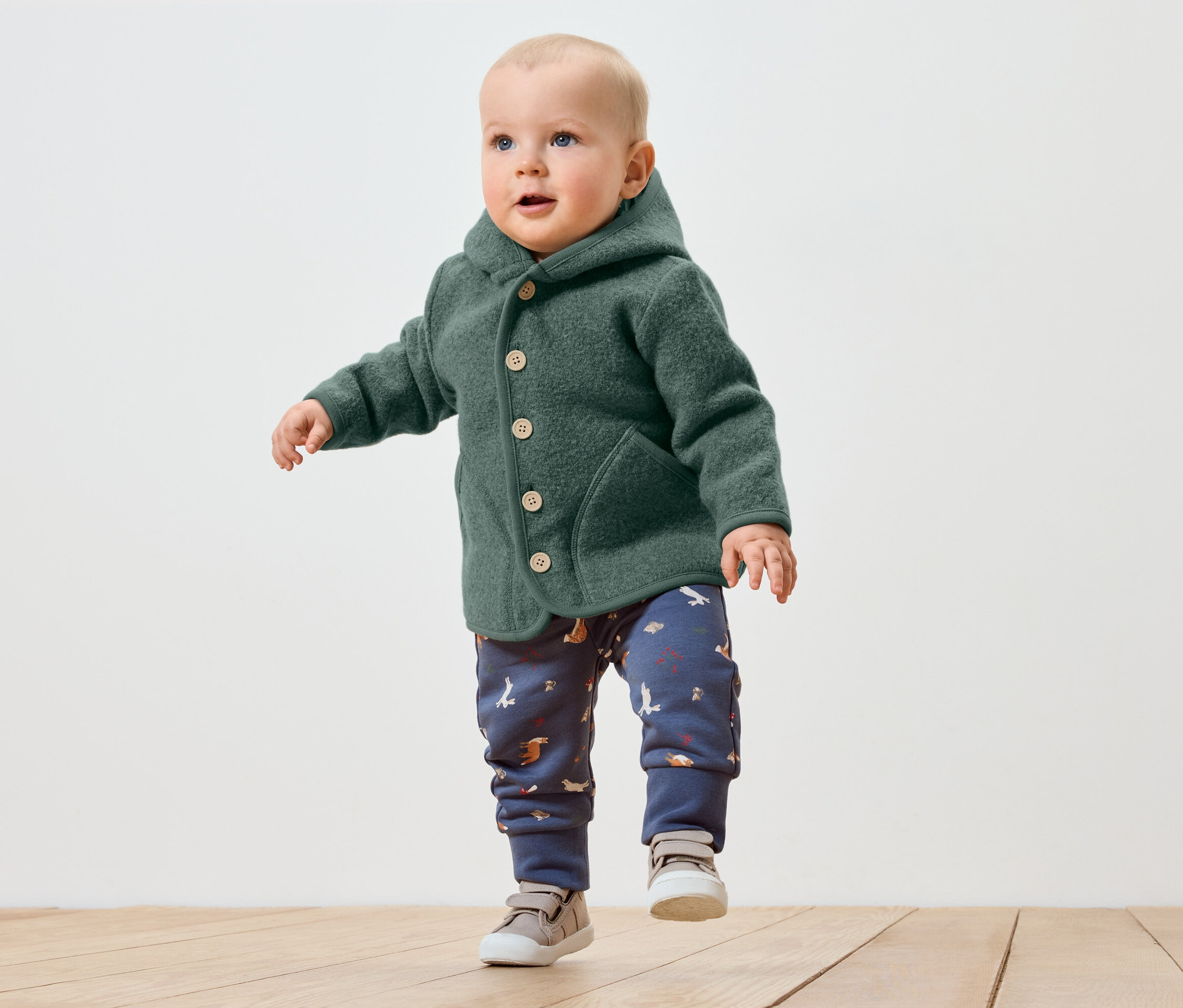 Batole v zeleném kabátku s kapucí a vzorovaných kalhotách dělá první krůčky po dřevěné podlaze.