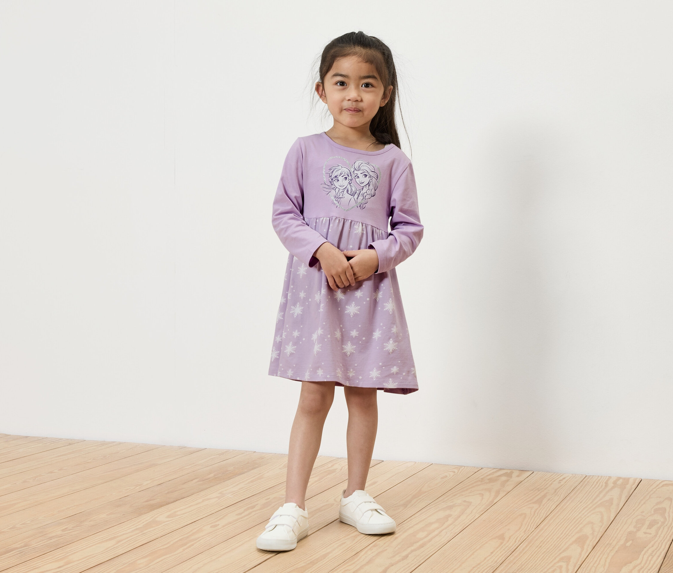 Dívka stojí a má na sobě světle fialové šaty s dlouhým rukávem s obrázkem Elsy a Anny a bílé tenisky.
