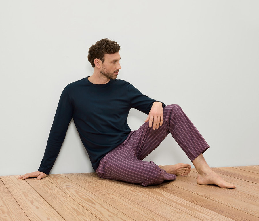 Muž sedí na dřevěné podlaze, opírá se o bílou zeď, má na sobě tmavě modré tričko s dlouhým rukávem a fialové pruhované kalhoty.