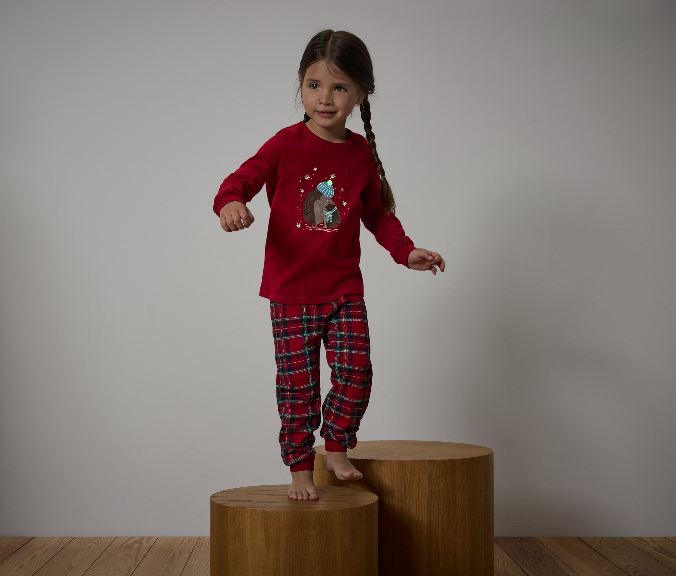 Dívka v červeném pyžamu s kostkovanými kalhotami stojí na dřevěných podstavcích.