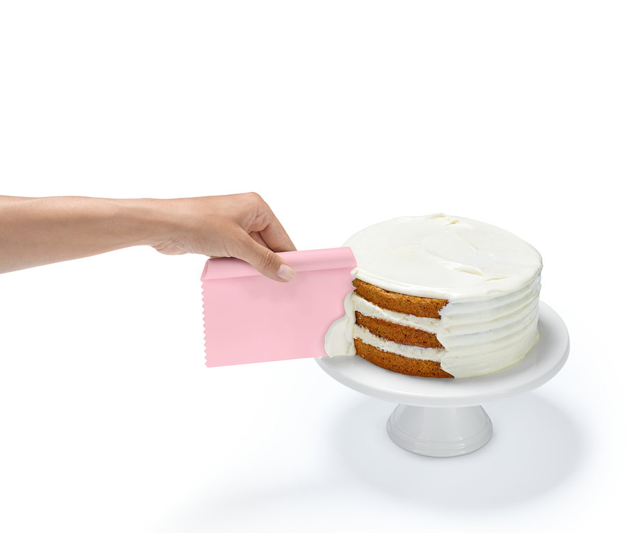 Ruka nanáší polevu na třívrstvý dort s růžovou špachtlí, který je umístěn na bílém podstavci.