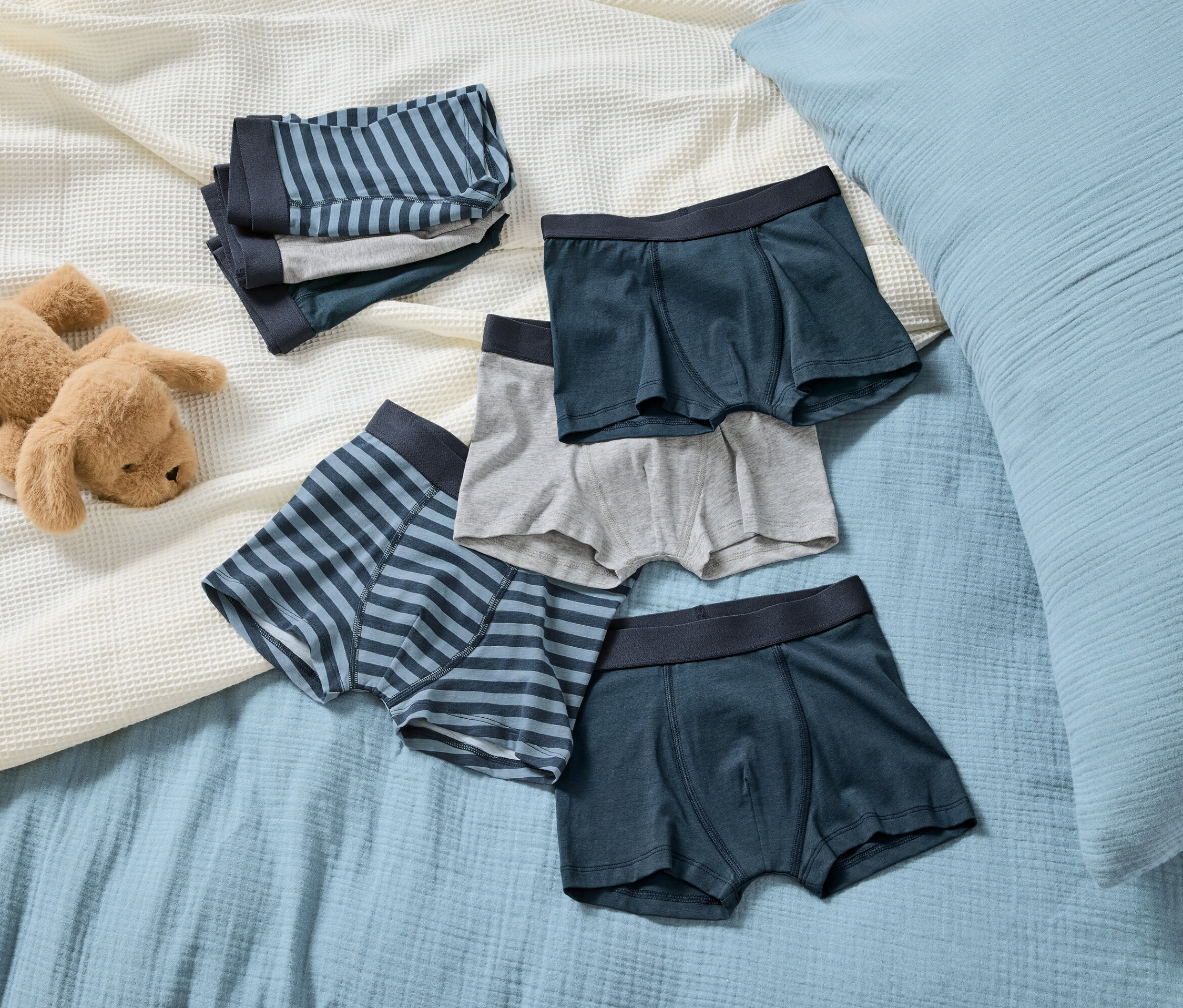Na světle modré dece leží hnědý plyšový pes vedle hromádky pruhovaných a hladkých chlapeckých boxerek.