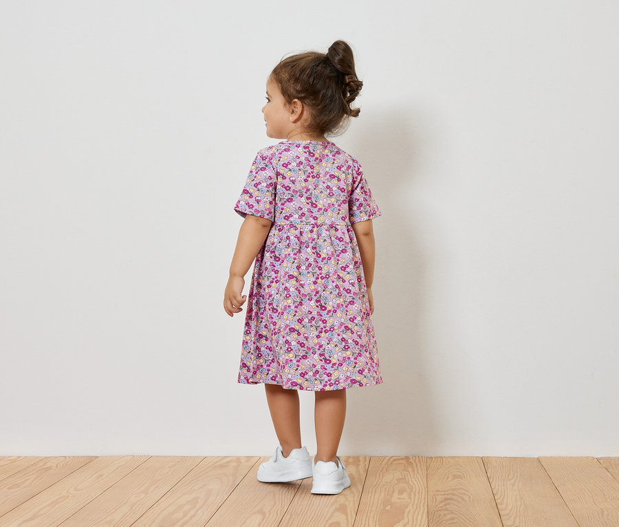 Holčička v šatech s květinovým vzorem a bílých teniskách stojí zády ke kameře.