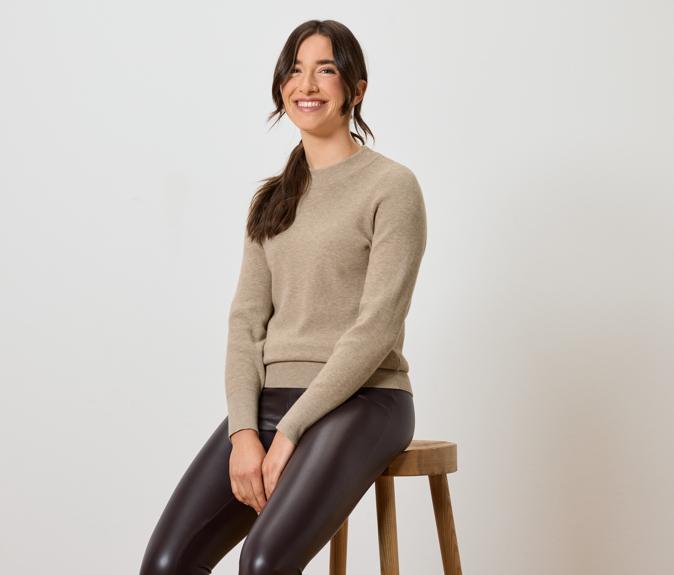 Žena sedí na stoličce v hnědých kožených legínách a béžovém svetru.