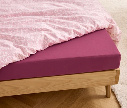 Detail dřevěné postele s bobulovým žerzejovým napínacím prostěradlem standardní velikosti a bílo-růžovým povlečením.