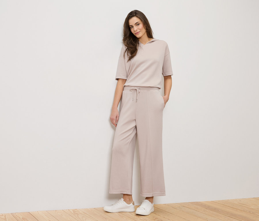 Žena stojí před bílou stěnou a má na sobě béžový outfit a bílé tenisky.