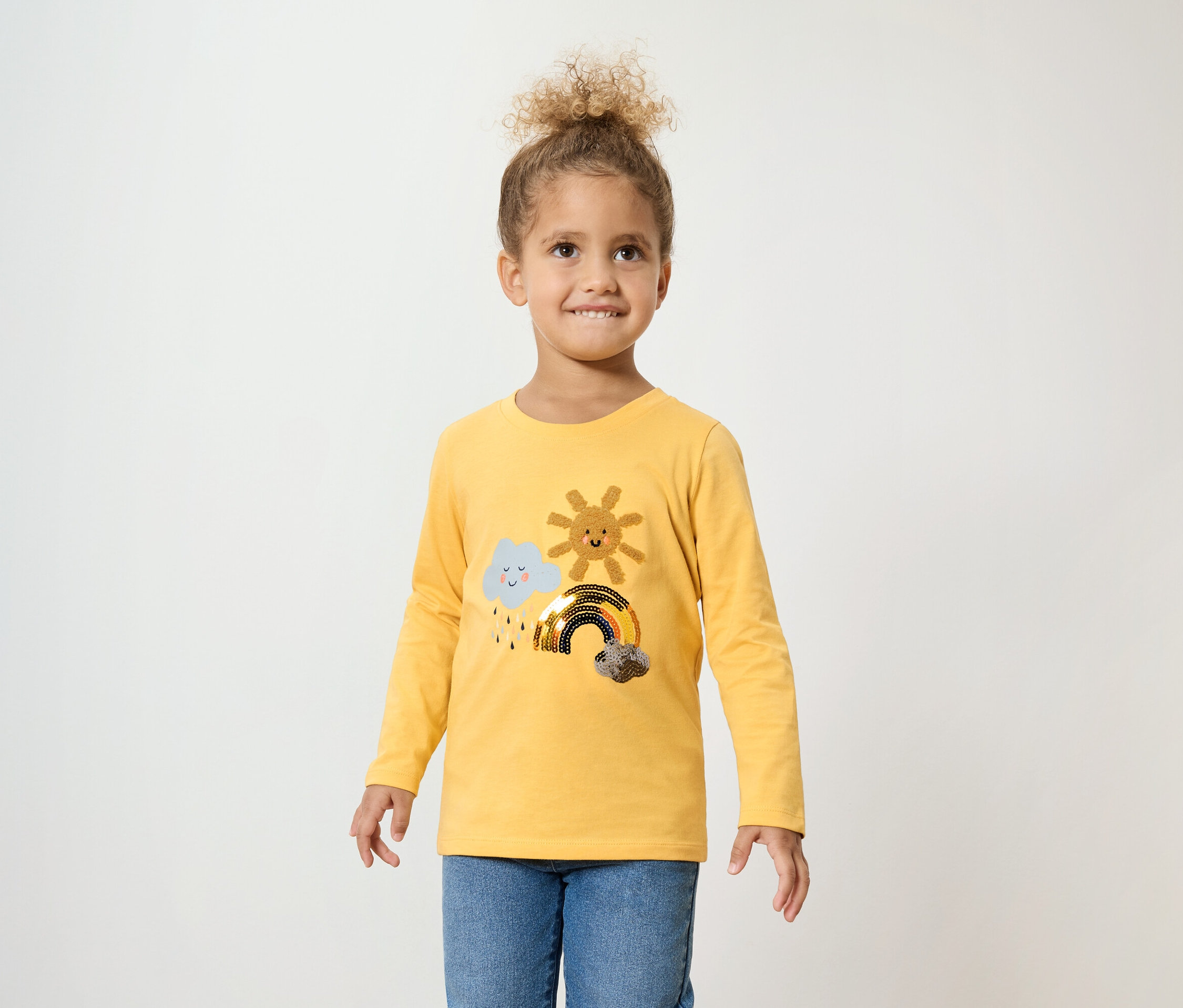 Dívka s kudrnatými vlasy má na sobě žluté tričko s dlouhým rukávem s obláčkem, sluncem a duhou a modré džíny.