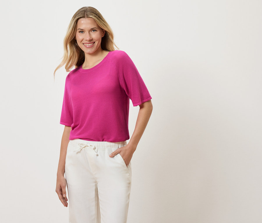 Žena s blond vlnitými vlasy má na sobě růžové tričko a bílé kalhoty a dává si ruku do kapsy.