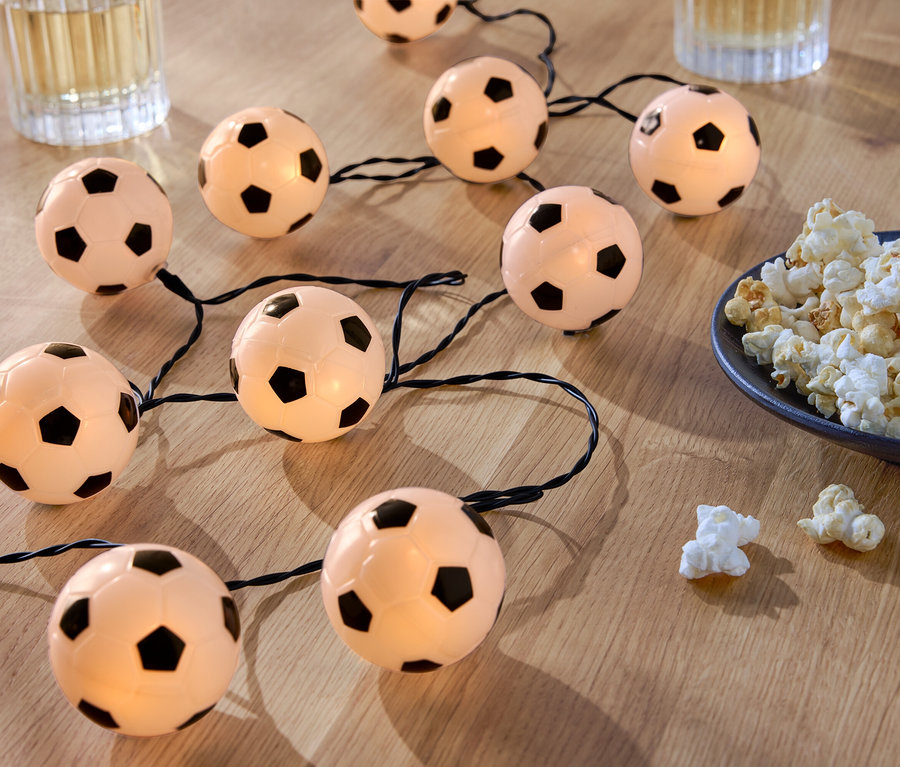 Světelný řetěz ve tvaru fotbalového míče leží na dřevěném stole vedle misky s popcornem a dvou sklenic s nápoji.