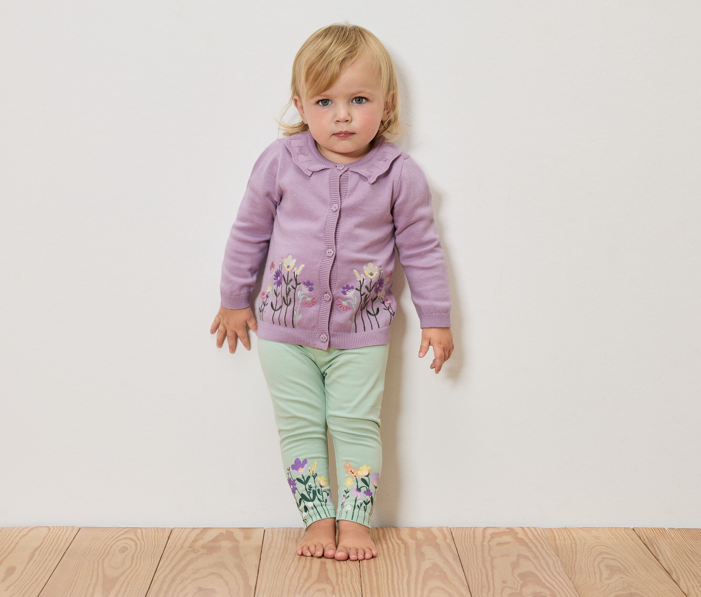 Blondýna stojí bosa před bílou stěnou, má na sobě fialový pletený svetr s květinovým vzorem a zelené legíny s květinami na nohavicích.