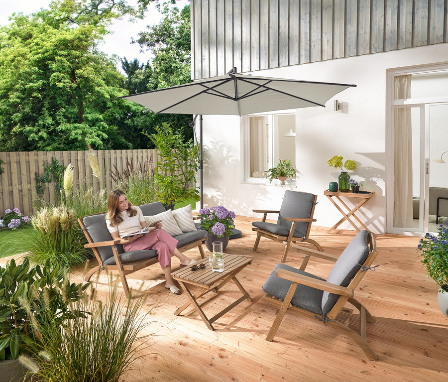 Žena sedí na skládací zahradní sedací soupravě s prémiovými polštáři, čte si a je obklopena zahradním odkládacím stolkem a 2 venkovními polštáři s lněným omakem.