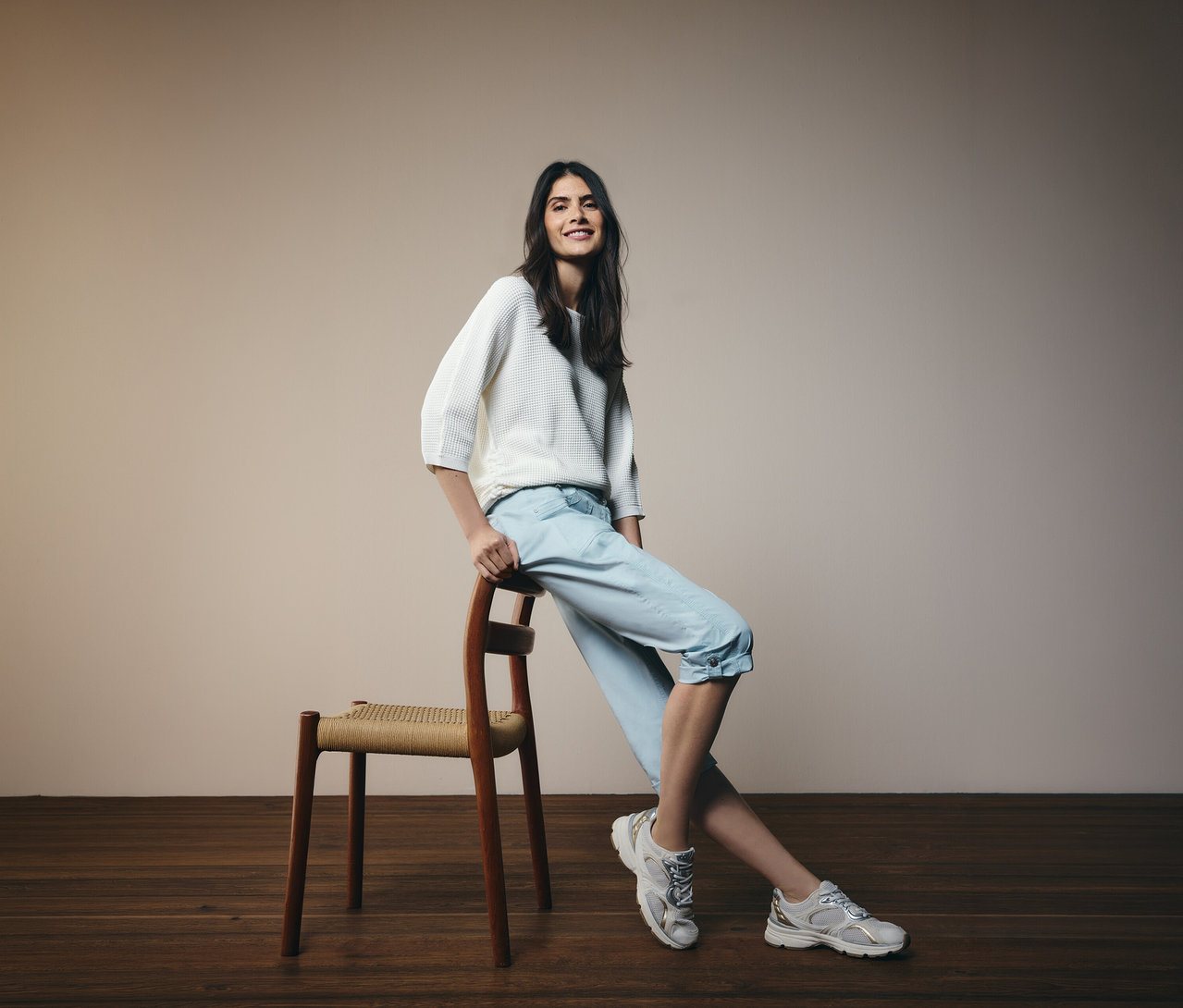 Žena se opírá o židli a má na sobě světle modré 3/4 kalhoty Cecil »New York« a bílý svetr.