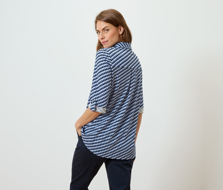 Žena s hnědými vlasy v modro-bílé vzorované blůze a tmavých kalhotách stojí zády k divákovi a dívá se přes rameno.