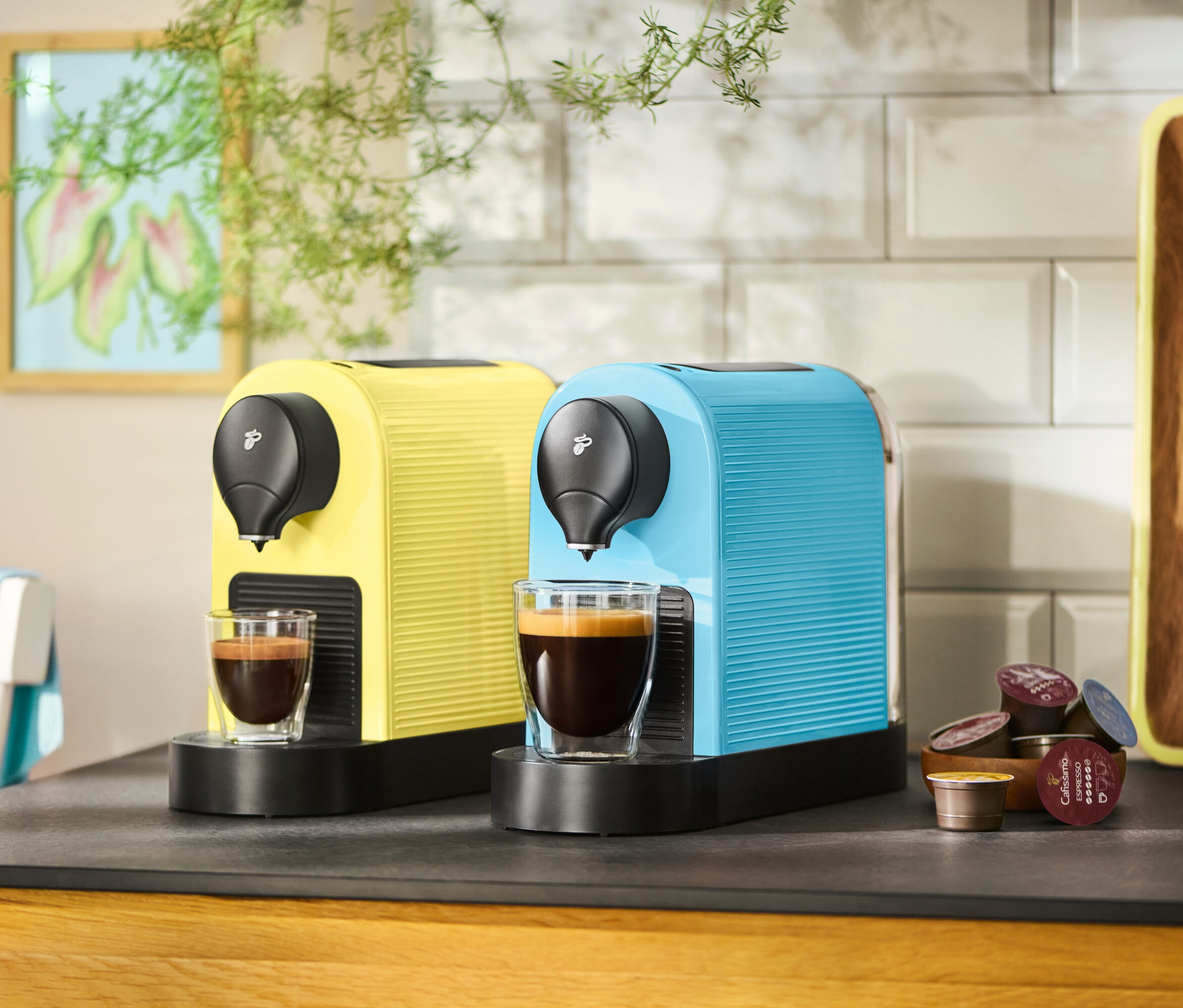 Dva kávovary ve žluté a modré barvě stojí na kuchyňské lince se dvěma šálky kávy a kávovými kapslemi.