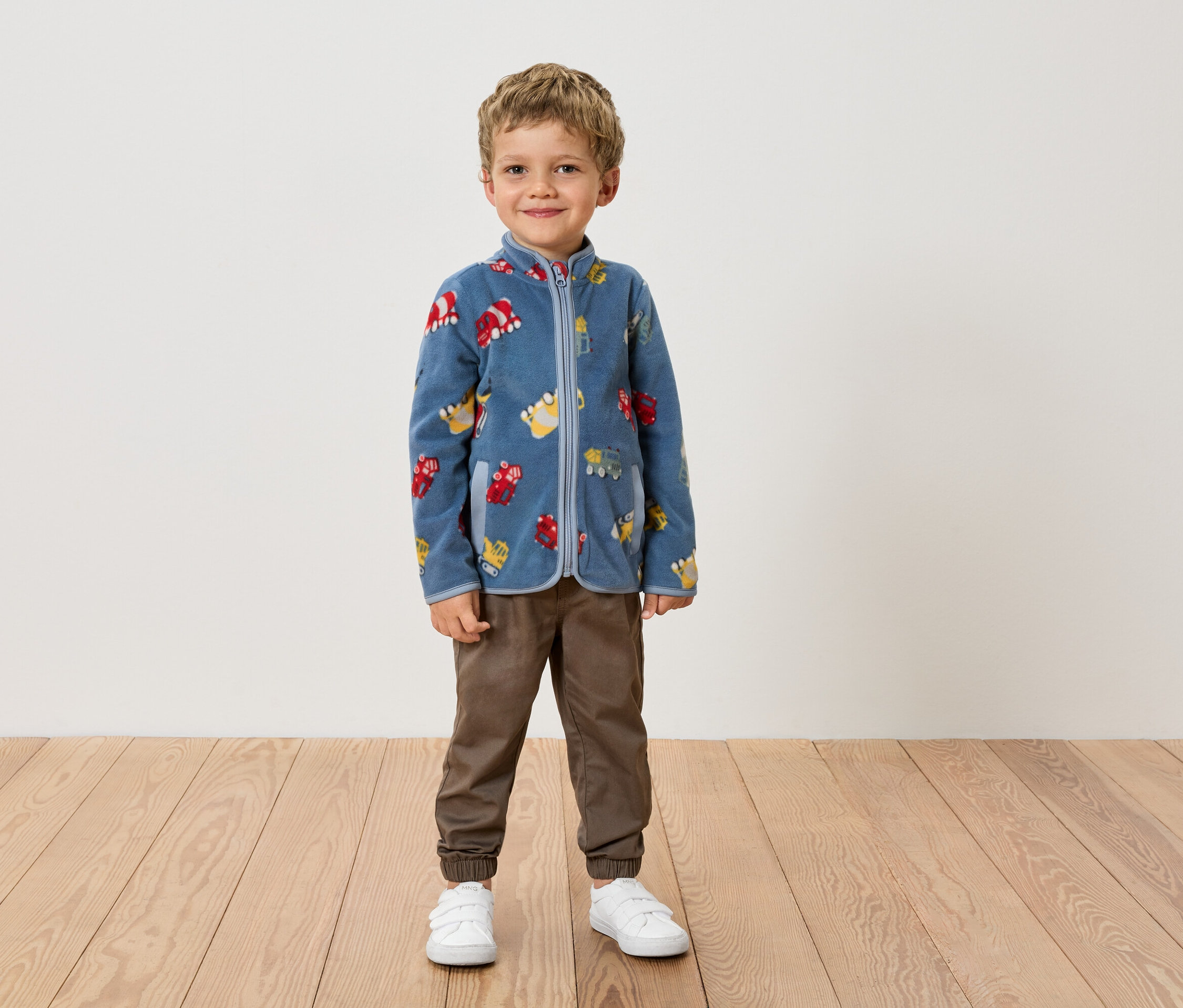 Chlapec stojí u zdi v modré fleecové bundě se vzorem vozidel, hnědých kalhotách a bílých botách.