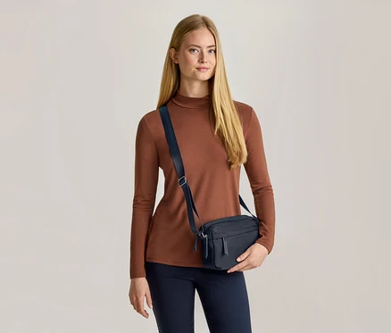 Žena s dlouhými blond vlasy má na sobě hnědý top s dlouhým rukávem, tmavě modré kalhoty a tmavě modrou crossbody tašku. Tašku drží levou rukou a dívá se dopředu.