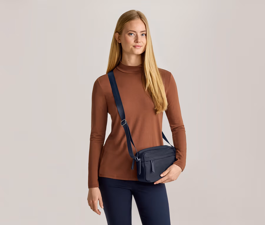 Žena s dlouhými blond vlasy má na sobě hnědý top s dlouhým rukávem, tmavě modré kalhoty a tmavě modrou crossbody tašku. Tašku drží levou rukou a dívá se dopředu.