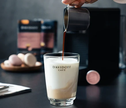 Ruka nalévá kávu do sklenice s mlékem. V pozadí je kávovar a balení DAVIDOFF CAFÉ Crema Intense.