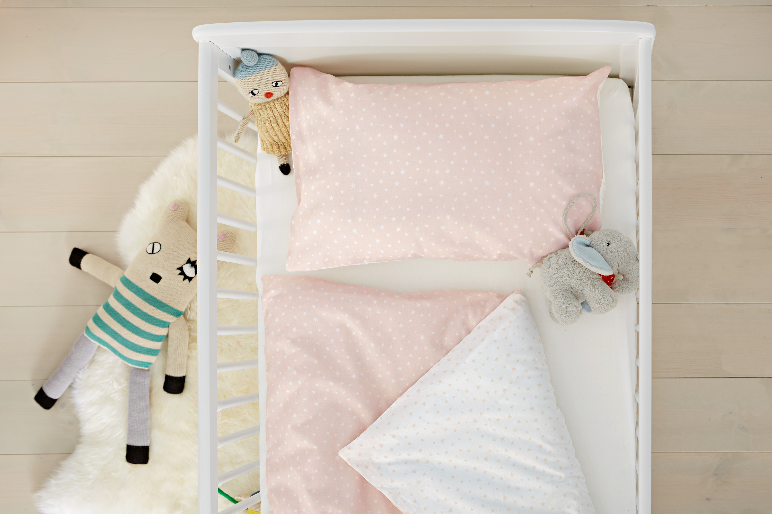 Pohled shora na bílou postýlku s růžovým povlečením s hvězdičkami, plyšovou hračkou a dekou.