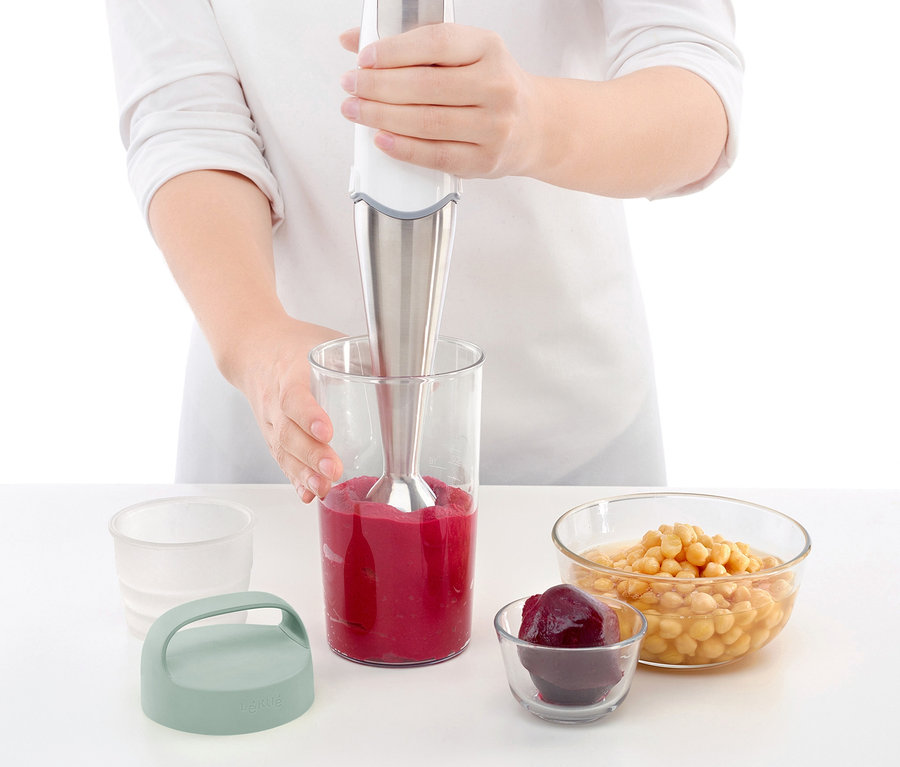 Žena mixuje smoothie v mixovací nádobě. Vedle stojí cestovní dóza na oběd a misky s ingrediencemi.