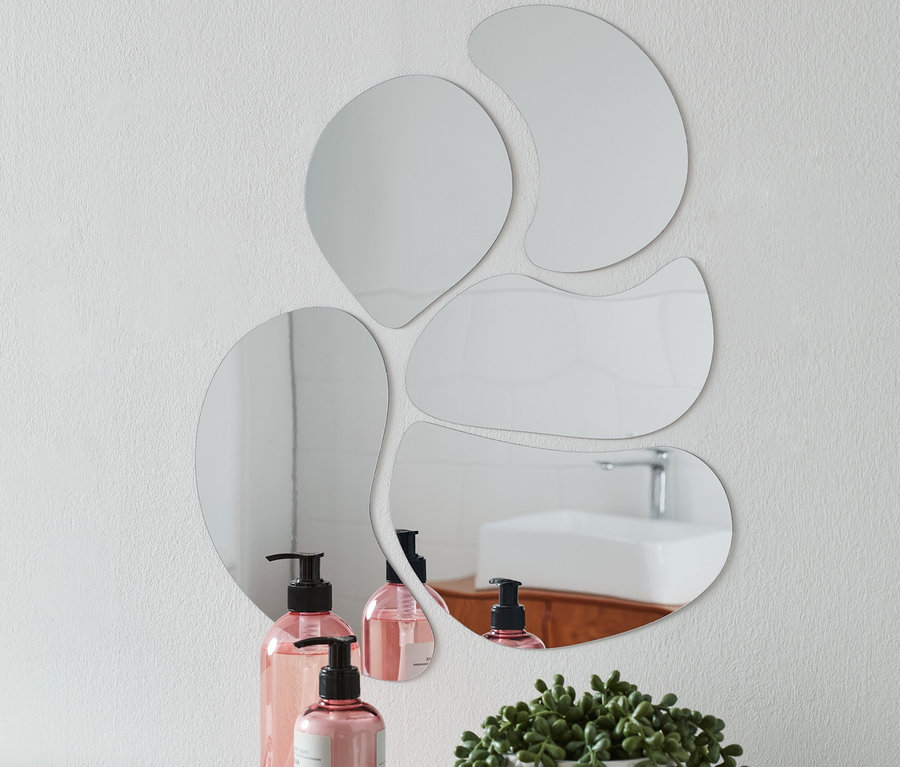 Na bílé zdi je pětice samolepicích zrcadlových nálepek různých tvarů. Na bílé polici pod nimi stojí dvě růžové lahve s pumpičkou, podnos s šperky a květina v bílém květináči.