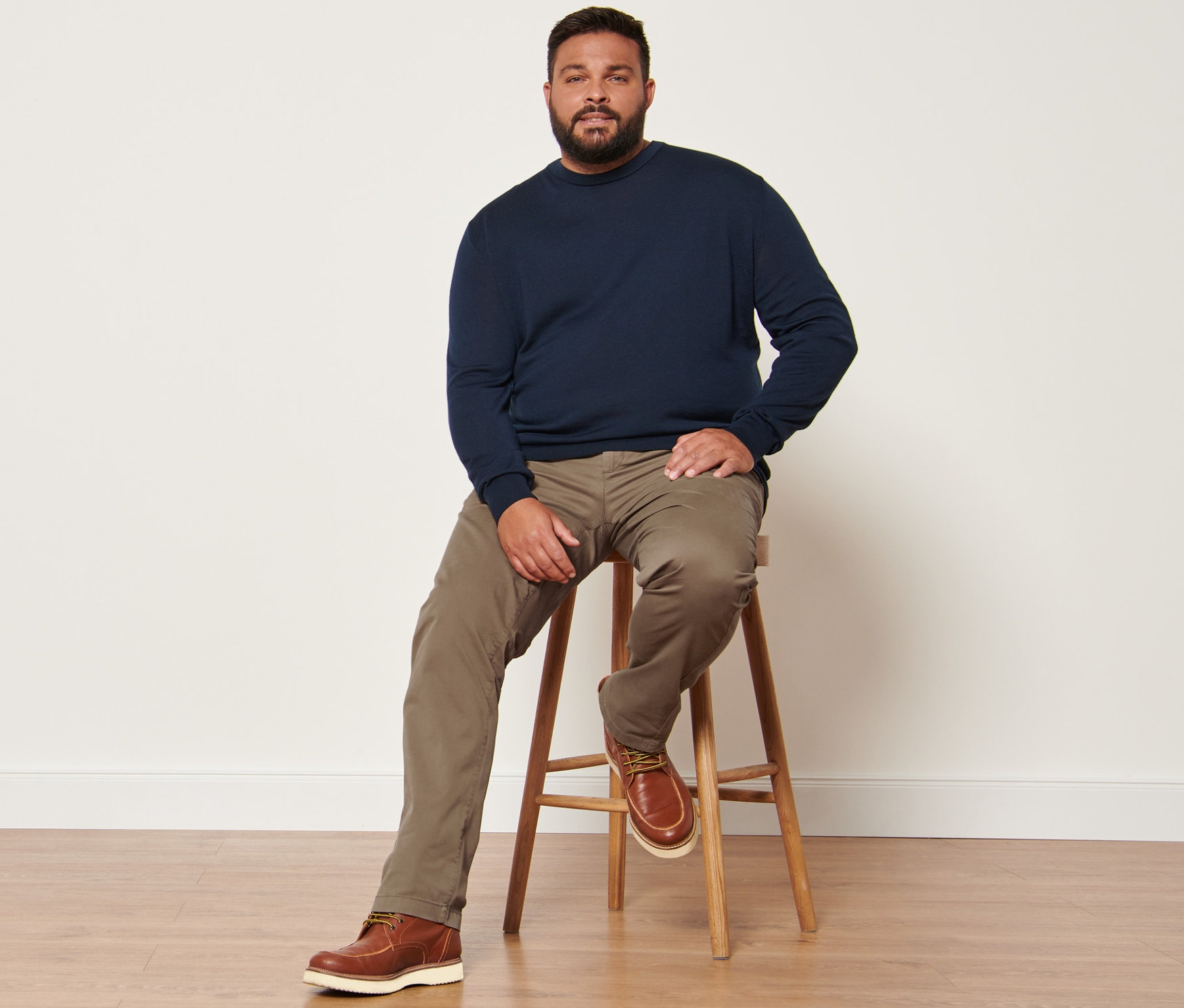 Muž sedí na stoličce a má na sobě námořnicky modrý svetr s vlnou merino a pískové kalhoty chino.