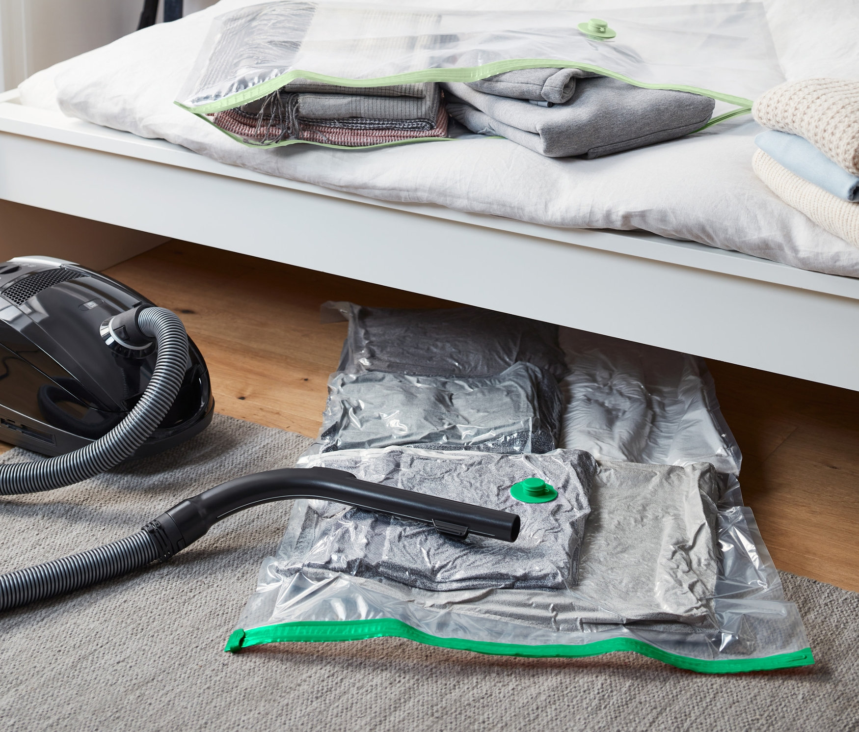 Černé vakuum stojí vedle postele. Na posteli a pod ní leží vakuové úložné pytle, 2 ks, horizontální.