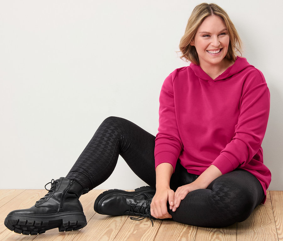 Žena sedí na dřevěné podlaze a má na sobě růžovou mikinu s kapucí z modalové směsi a černé termolegíny s vyraženým vzorem.