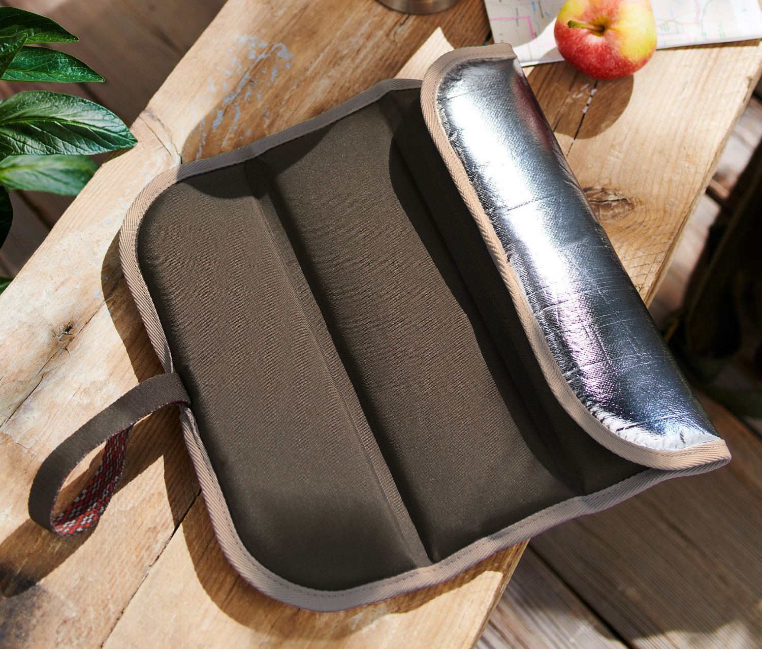 Skládací termo polštářek na sezení leží na dřevěném stole vedle termosky, jablka a batohu.