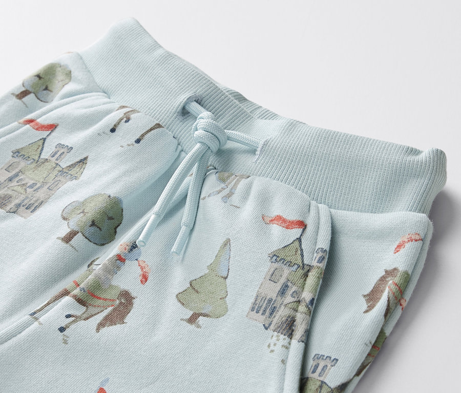 Detail světle modrých tepláků pro miminka s potiskem rytířů, hradů a stromů.