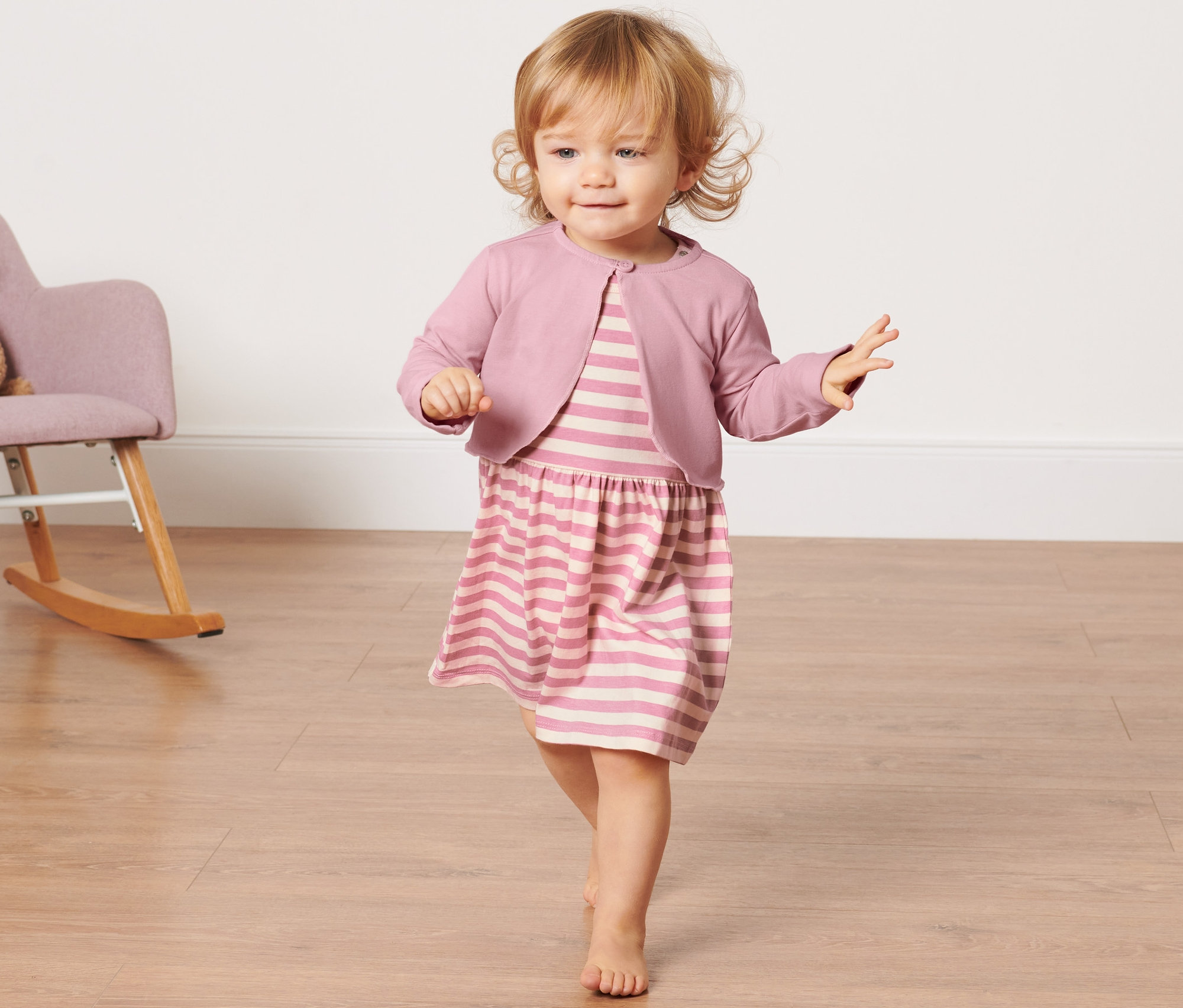Batole v pruhovaných šatech a bolerku kráčí po dřevěné podlaze. V pozadí se nachází houpací křeslo s plyšovým medvídkem.