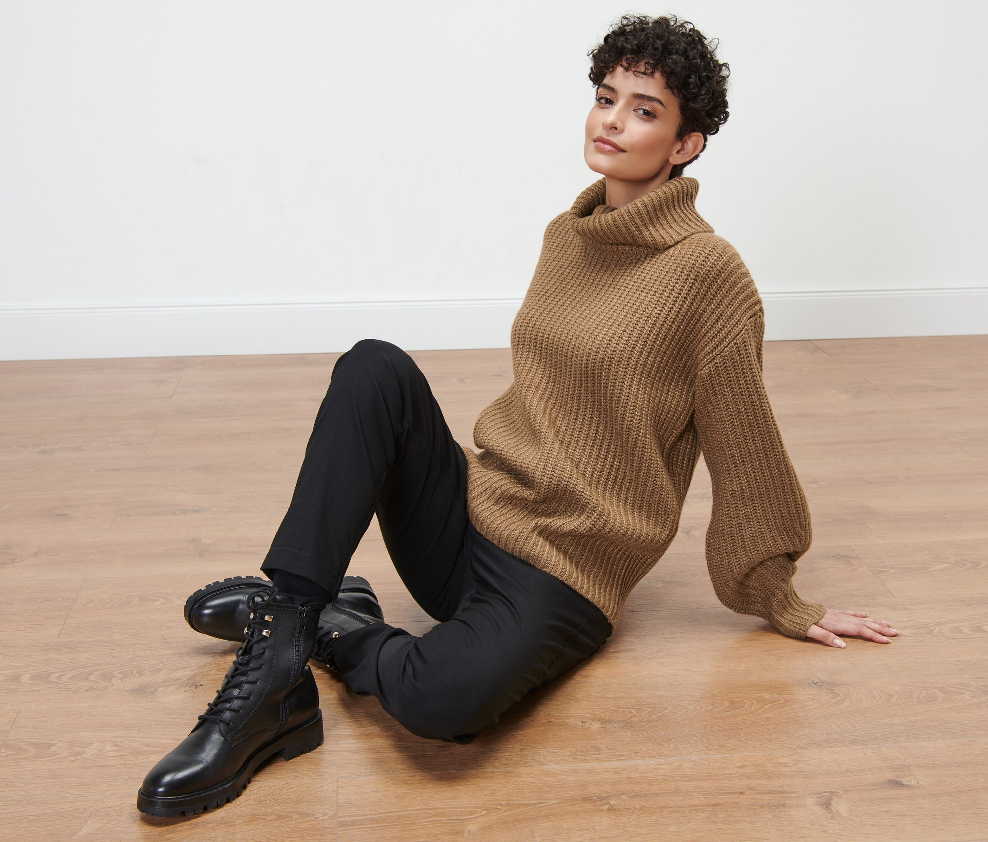 Žena sedí na podlaze a má na sobě hnědý svetr z hrubé pleteniny s rolákovým límcem, černé kalhoty a černé boty.