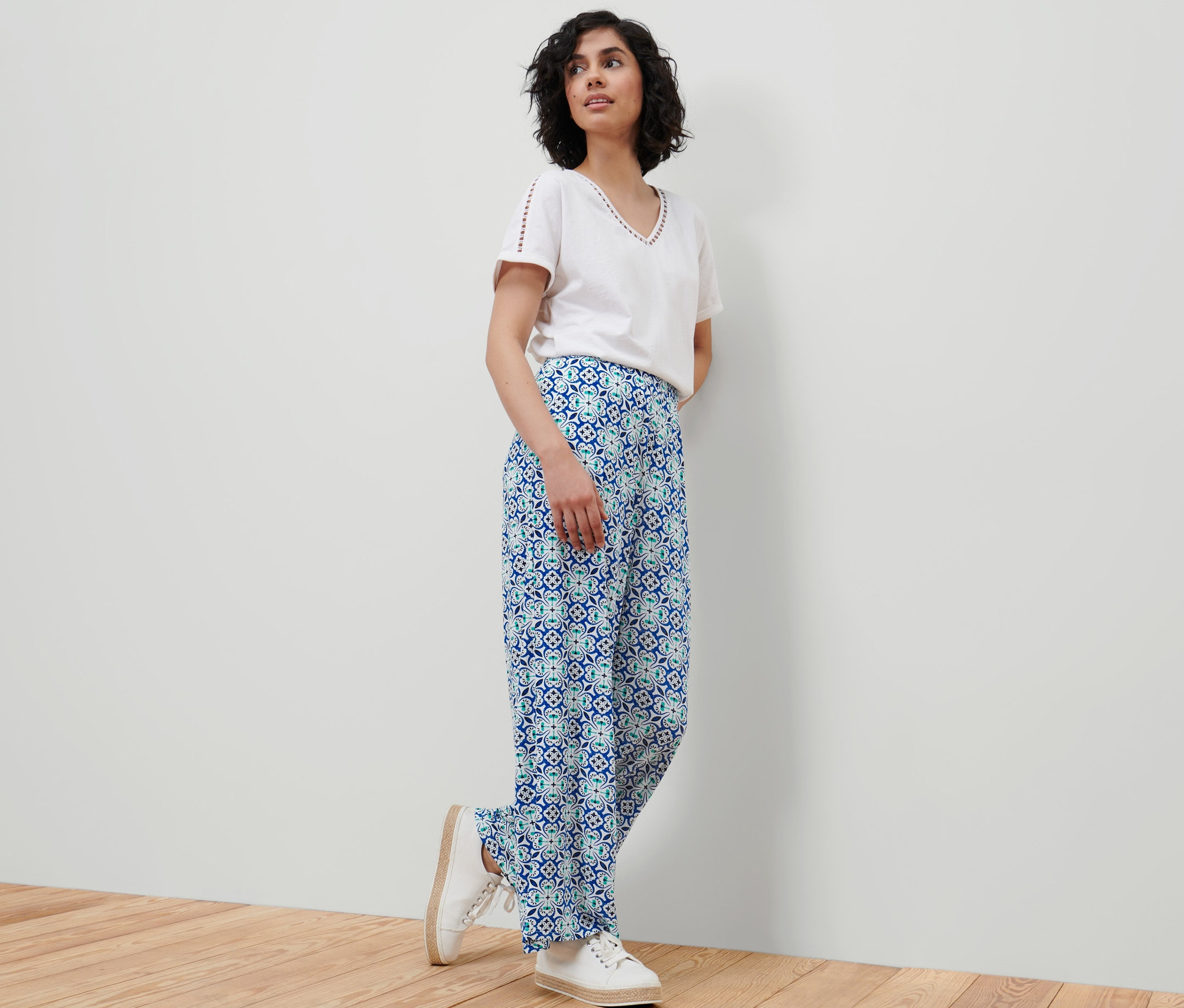 Žena stojí a opírá se o zeď a má na sobě bílé tričko a kalhoty se širokými nohavicemi s celoplošným potiskem.