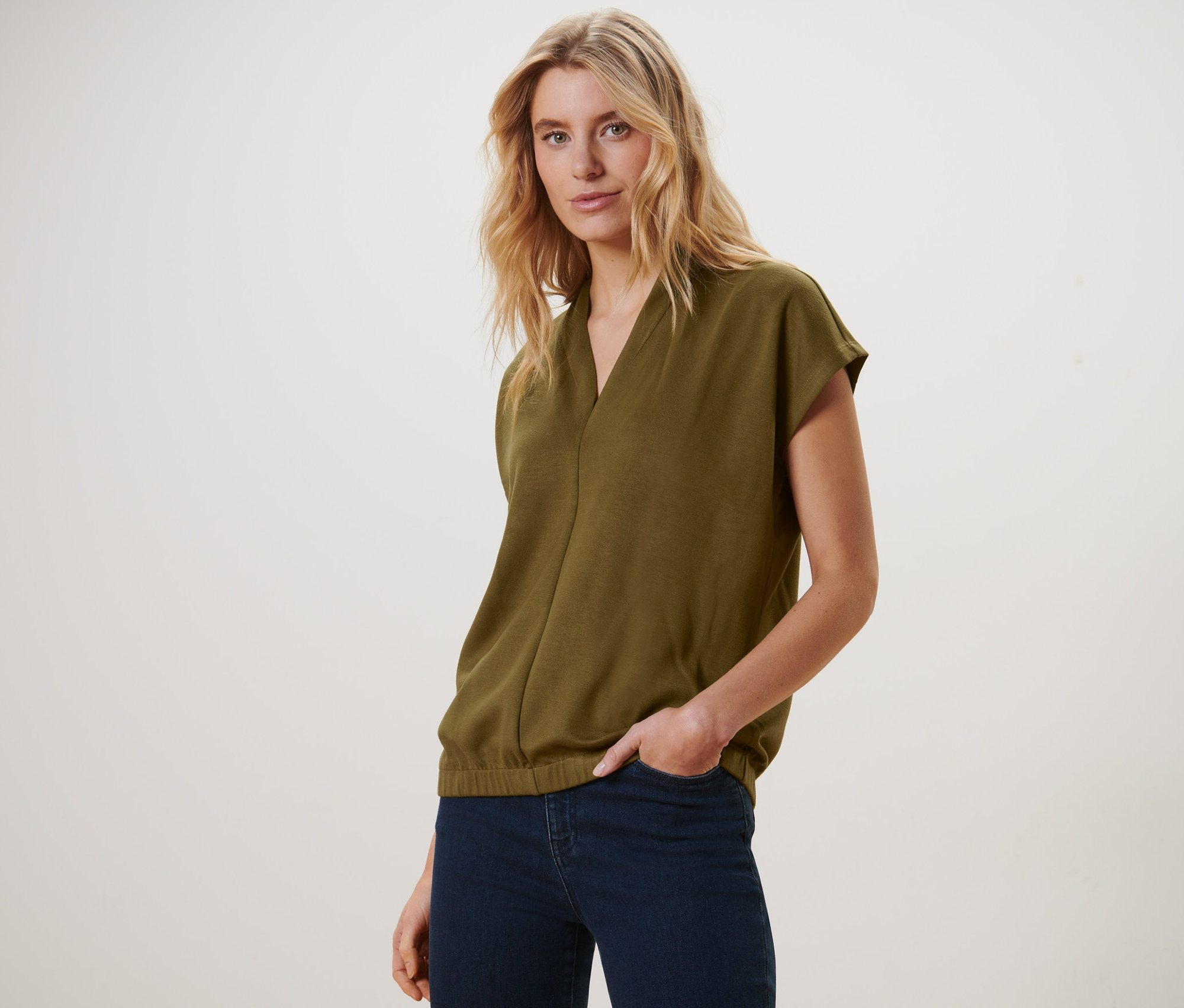 Blonďatá žena pózuje v olivově zeleném halénkovém tričku s výstřihem do V a tmavě modrých džínách.