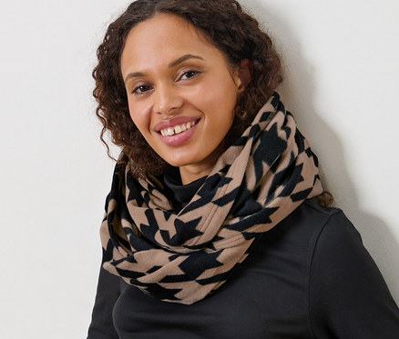 Žena s hnědými kudrnatými vlasy sedí a má na sobě fleecovou kruhovou šálu s potiskem kohoutí stopy.