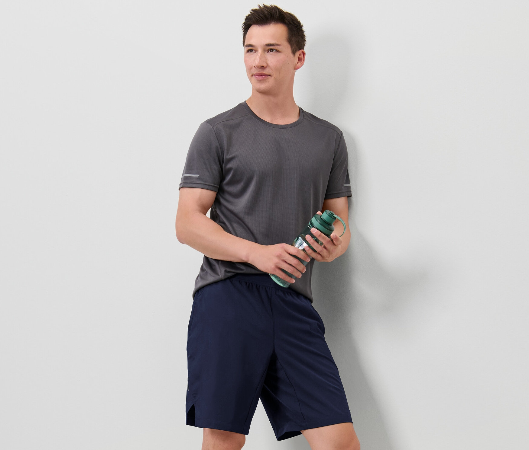 Muž v šedém tričku drží zelenou láhev s vodou a má na sobě sportovní šortky.