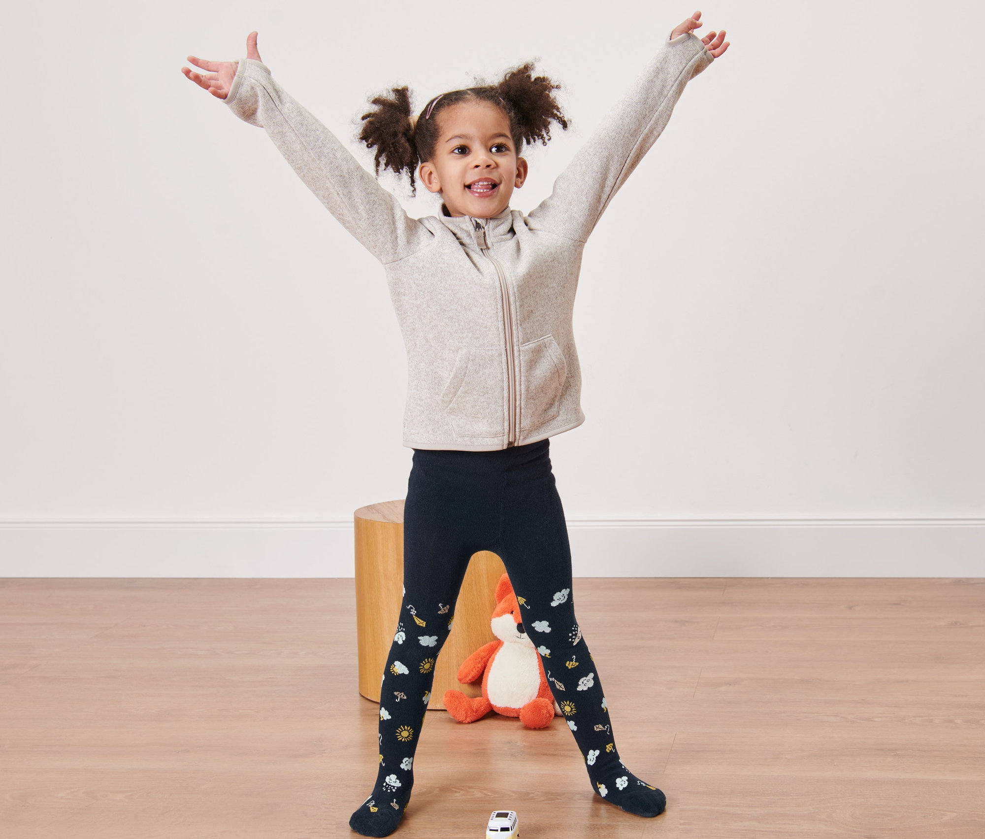 Usmívající se holčička stojí s rozpaženýma rukama a má na sobě modré punčocháče s potiskem.