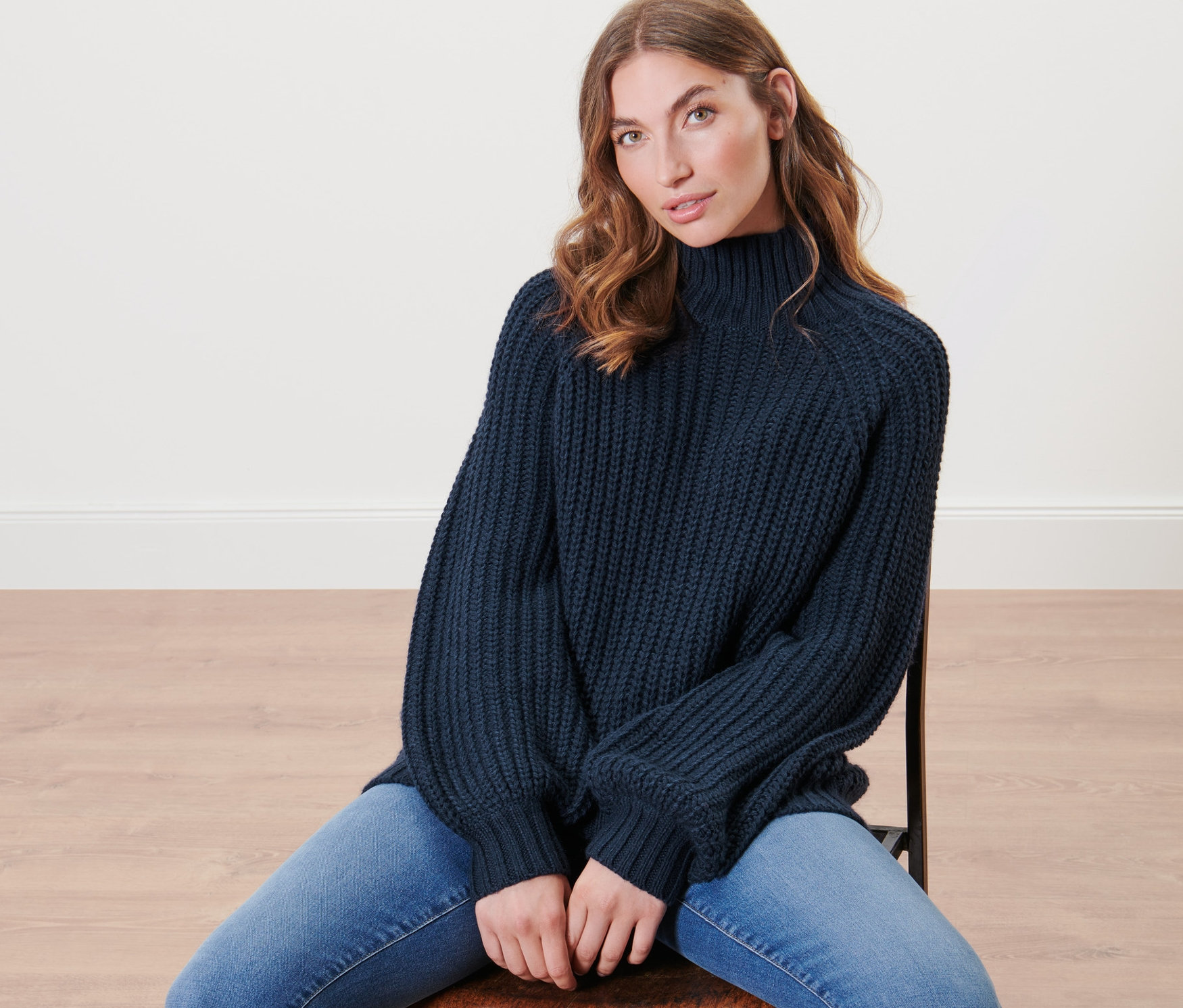 Žena sedí na židli a má na sobě tmavě modrý pletený svetr se stojáčkem.