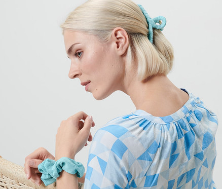 Blonďatá žena sedí v křesle, na zápěstí má gumičku do vlasů a ve vlasech sponu. Sada vlasových doplňků.