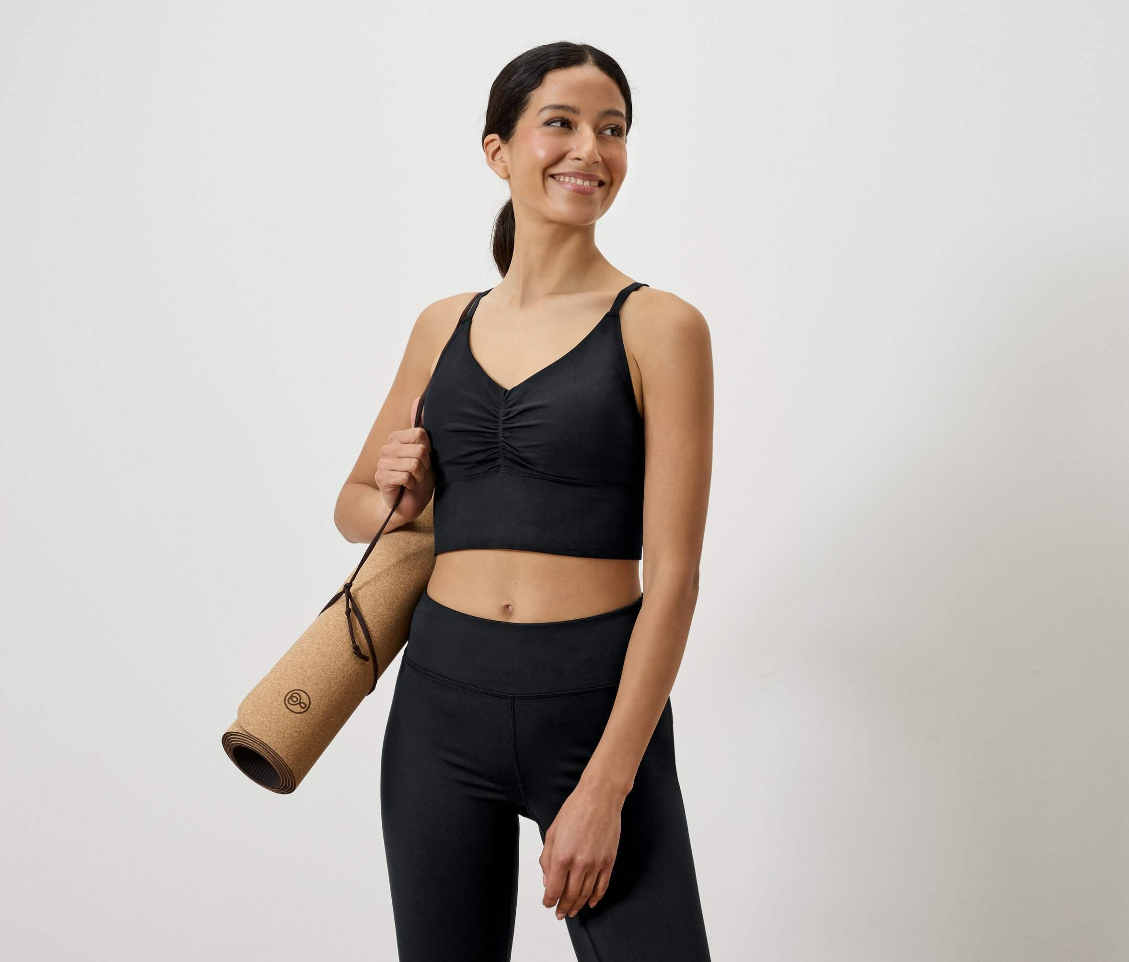 Usmívající se žena v černém sportovním oblečení drží pod paží srolovanou jógamatku.