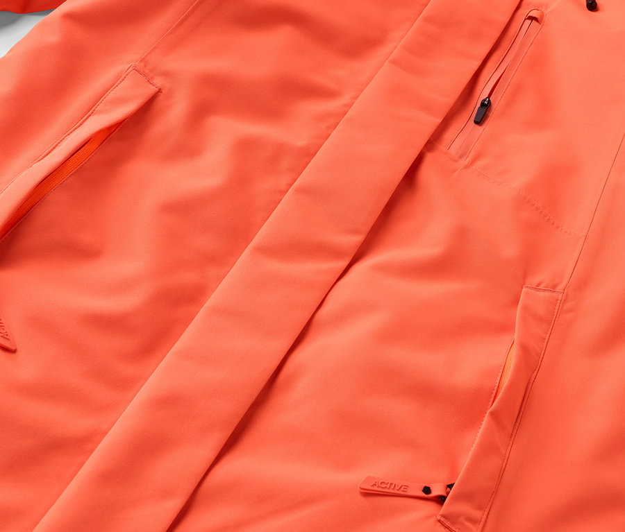 Detail oranžové bundy s kapsami a zipem.