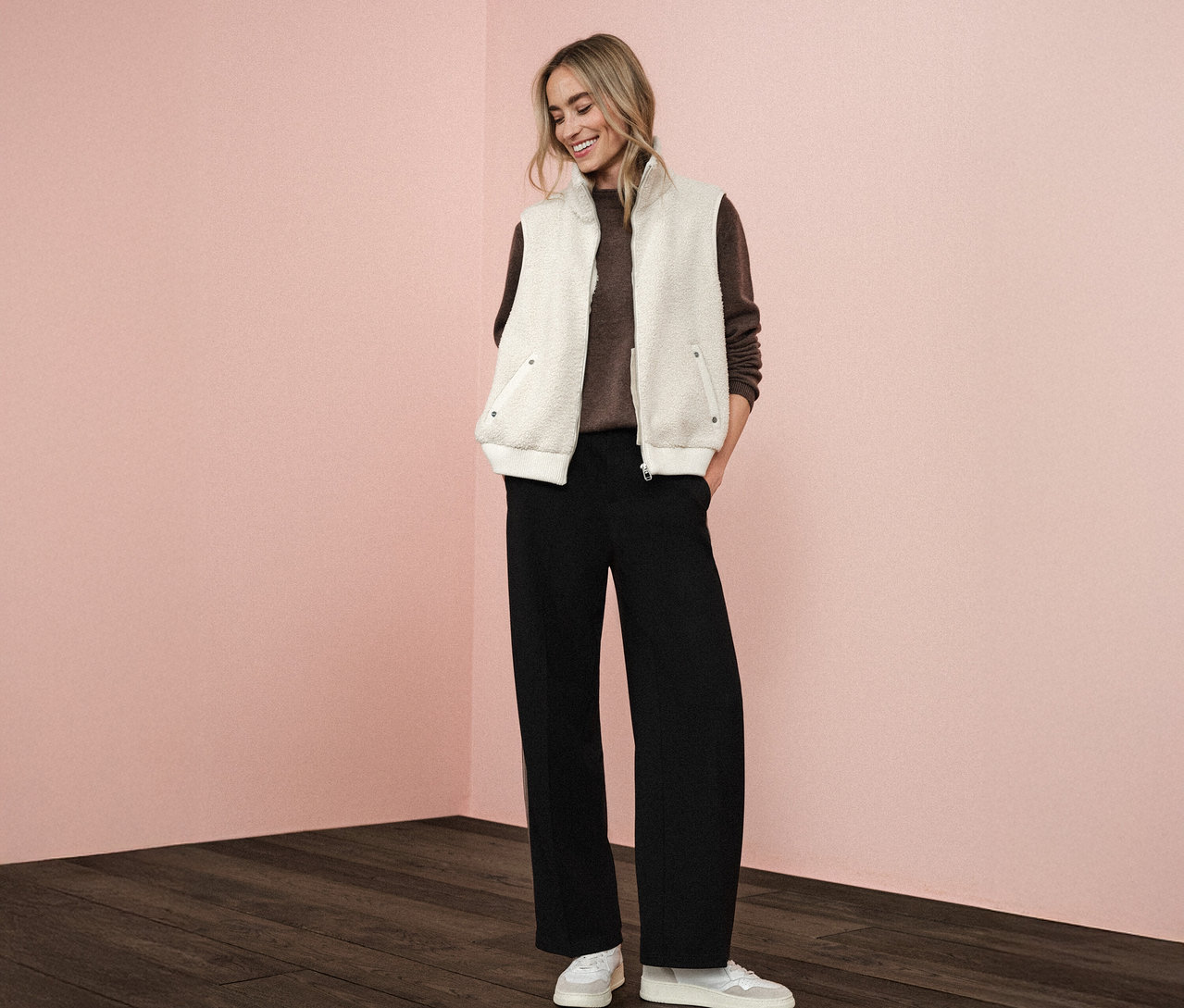 Žena má na sobě plyšovou vestu Street One přes hnědý svetr, v kombinaci s černými kalhotami a bílými teniskami.