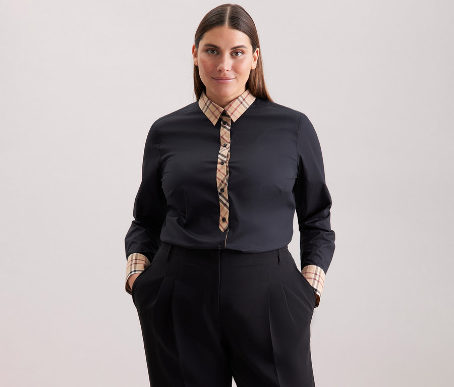 Žena s hnědými vlasy má na sobě černou popelínovou halenku Seidensticker s kostkovaným detailem a černé kalhoty.