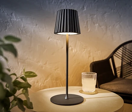 Černá dobíjecí LED stolní lampa stojí na stole vedle sklenice s pitím.